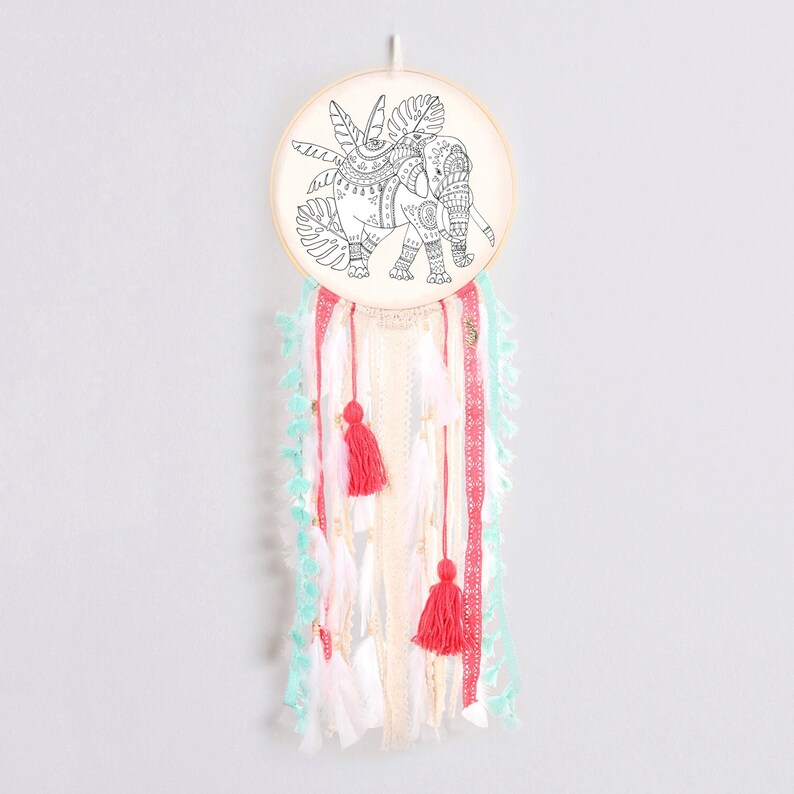 May include: A white wooden hoop with a black and white line drawing of an elephant with floral designs. The hoop is hanging from a white string and has white, pink, and turquoise tassels hanging from it.