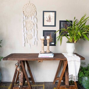 May include: A wooden table with two sawhorse legs, a white runner, and a stack of books with the title "Arte moderno". There are two candlesticks with white candles on the table, and a white dreamcatcher hanging on the wall behind the table.
