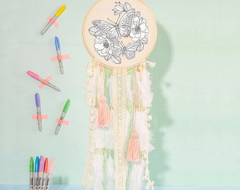 May include: A white dreamcatcher with a wooden hoop and a black and white butterfly design. The dreamcatcher has white feathers and pink tassels. The dreamcatcher is surrounded by colorful markers.