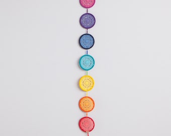 May include: A colorful chakra wall hanging featuring seven crocheted circular medallions in a rainbow of colors: red, orange, yellow, turquoise, blue, purple, and pink. Each medallion is connected by a string with beads and a tassel at the bottom.