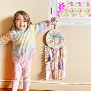 May include: A pastel rainbow colored knitted sweater with a young person wearing it. The person is standing in front of a wall with a unicorn dream catcher hanging on it. The dream catcher has white, pink, and purple feathers.