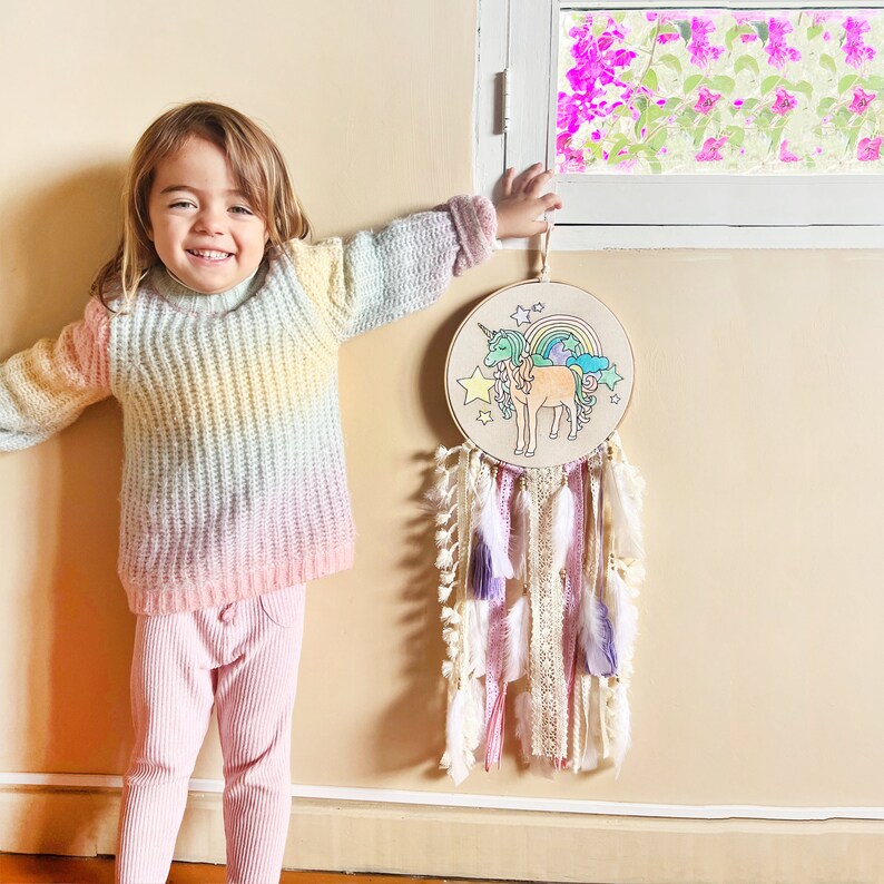 May include: A pastel rainbow colored knitted sweater with a young person wearing it. The person is standing in front of a wall with a unicorn dream catcher hanging on it. The dream catcher has white, pink, and purple feathers.