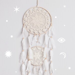 May include: A white dream catcher with two circular crocheted hoops and white feathers hanging from the bottom. The dream catcher is hanging on a light gray wall with white star and moon shapes.