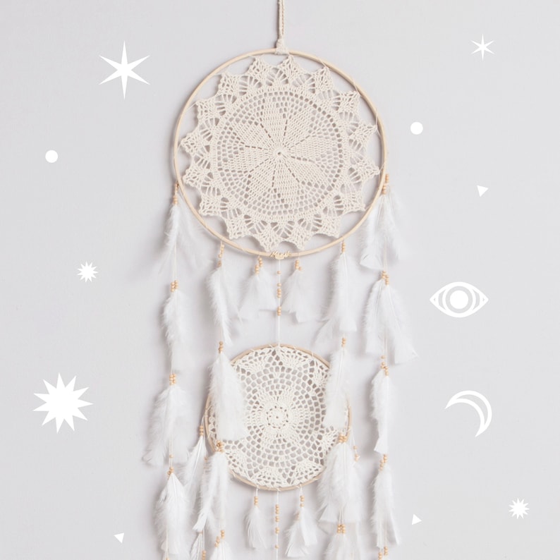 May include: A white dream catcher with two circular crocheted hoops and white feathers hanging from the bottom. The dream catcher is hanging on a light gray wall with white star and moon shapes.