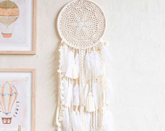 Puede incluir: Un tapiz de macramé color crema con un centro circular de ganchillo que presenta un complejo patrón geométrico. La pieza está adornada con múltiples hilos colgantes de flecos blancos, plumas suaves y pequeñas cuentas de madera texturizadas.