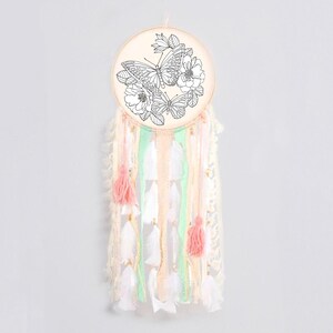 May include: A white dream catcher with a wooden hoop featuring a black and white line drawing of butterflies and flowers. The dream catcher has pink and green ribbons and white feathers.