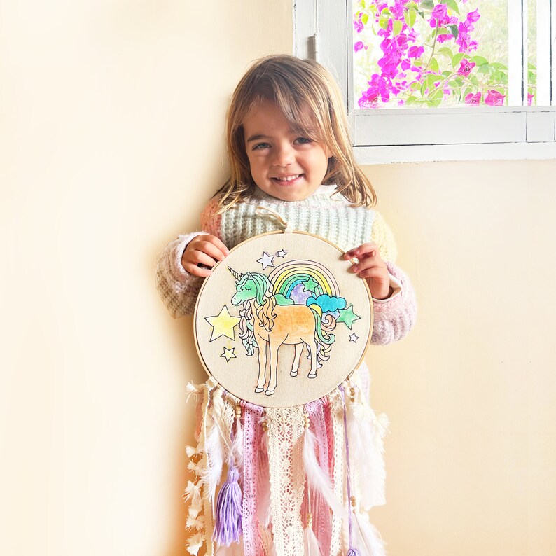 May include: A young person holds a wooden hoop with a colorful unicorn design and rainbow. The hoop is decorated with white feathers and pink and purple yarn.