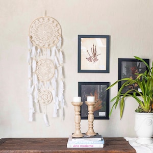 May include: A cream-colored dreamcatcher with three circular woven designs and white feathers hangs on a wall. Two framed botanical prints and two wooden candle holders with white candles sit on a wooden table, with a potted plant.