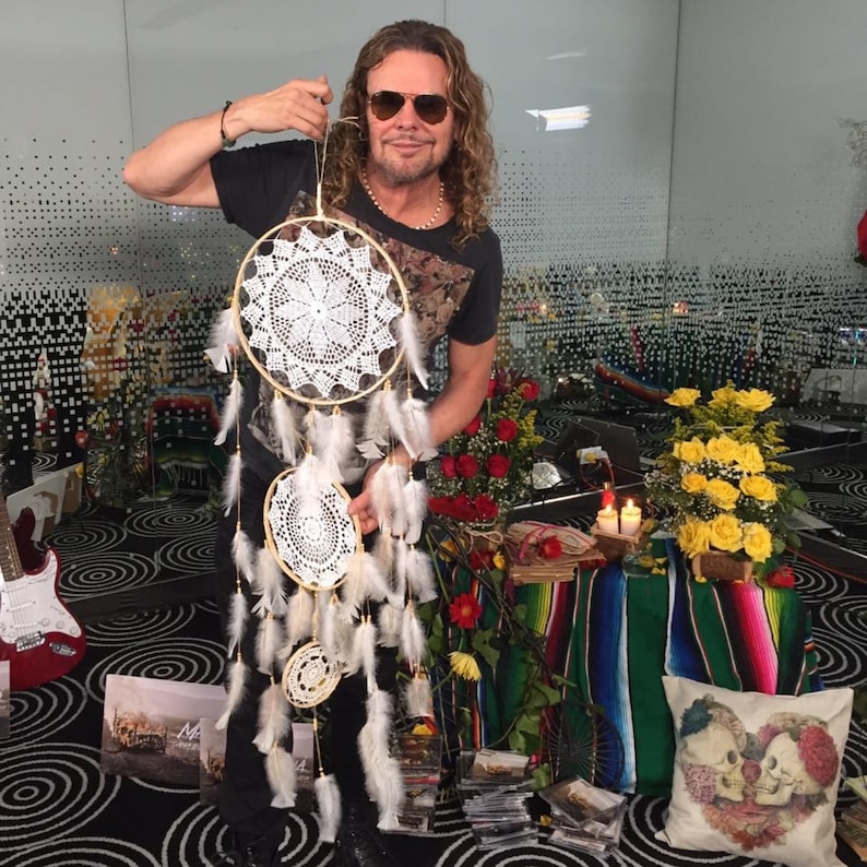 May include: A man wearing sunglasses holds a white dreamcatcher with three hoops and white feathers. The dreamcatcher is hanging from a wooden dowel. The man is standing in front of a colorful background with flowers and candles.