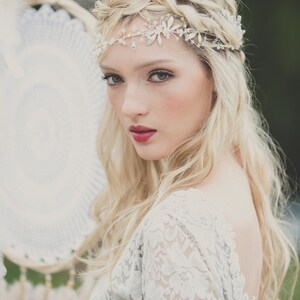 May include: A woman with long blonde hair wearing a white lace dress and a silver headband with rhinestones. She is looking at the camera with a soft smile.