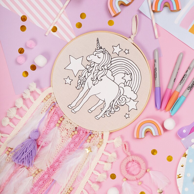 May include: A white unicorn with a rainbow and stars on a beige embroidery hoop. The hoop is surrounded by pink and white feathers, beads, and a purple tassel.