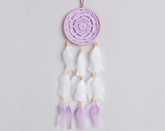 May include: A dream catcher featuring a circular frame wrapped in light purple yarn with a matching intricate crochet web center. Five hanging strands are adorned with small wooden beads, white feathers, and light purple feathers at the ends.