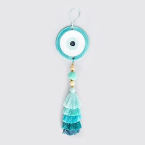 May include: A round, turquoise and white Evil Eye wall hanging with a tassel. The eye design features concentric circles of blue and white. Below the eye are wooden beads and a layered tassel in shades of blue.