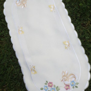 May include: Oval-shaped, off-white table runner with a scalloped edge. The runner features embroidered designs of butterflies, flowers in pink, blue, and yellow, and a bunny. The design is centered with a white stitched border.