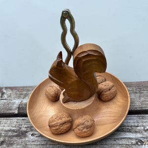 May include: A round wooden serving bowl featuring a central carved squirrel figure and a vertical metal nutcracker mechanism. Four whole walnuts are arranged on the surface of the bowl around the base of the squirrel figure.