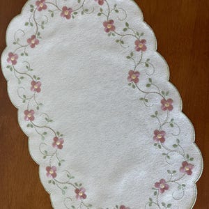 May include: Oval-shaped, off-white placemat with a scalloped edge. The placemat is embroidered with a floral design featuring pink flowers and green leaves. The flowers are arranged in a continuous pattern around the edge.