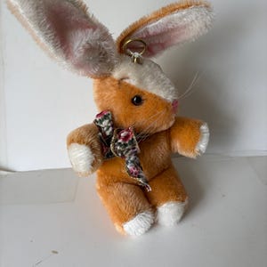 May include: A plush orange and white bunny rabbit with a floral bow tie. The bunny has long ears with pink and white fur, black eyes, and a gold ring attached to its head. The bunny's paws and face are white.