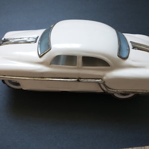 May include: A white toy car with silver-colored trim along the hood, roof, and sides. The vehicle features light blue tinted windows, a streamlined body shape, and white-walled tires visible beneath the silver side molding and bumpers.