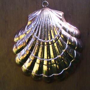 May include: A decorative, metallic seashell wall hanging. The shell is made of a shiny, copper-colored metal with a scalloped design. It has a loop at the top for hanging and reflects light, creating a warm, golden glow. The shell is approximately 12 inches wide.
