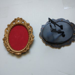 May include: Two ornate, oval picture frames. One is gold-colored with a red interior and a decorative border. The other is black with a decorative edge and a black easel back. The frames are empty.