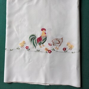 May include: White tablecloth with an embroidered design featuring a rooster, hen, and two chicks. The rooster has a red comb and a green tail. The hen is brown and the chicks are yellow. Red and yellow flowers are also embroidered.