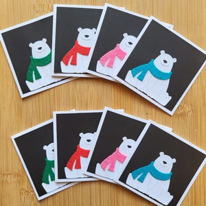 Peut inclure: Un ensemble de huit cartes de vœux carrées illustrant un ours polaire blanc sur fond noir. Chaque ours porte une écharpe texturée de couleur verte, rouge, rose ou bleu sarcelle. Les cartes sont disposées en deux rangées de quatre, mettant en valeur la variété des couleurs d'écharpes sur le design noir et blanc.