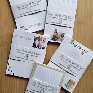 May include: A selection of notepads with different designs and text, such as "things to fetch," "Honey-Do List," and "Enjoy the little things." Each notepad is wrapped with a band featuring the text "Say It With A Card" and a website address.