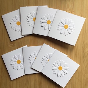 May include: A set of white square greeting cards, each embossed with a daisy design. The daisies have white petals and yellow centres. The cards are displayed on a light brown wooden surface.