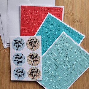 Peut inclure: Un ensemble de papeterie comprenant deux cartes rectangulaires avec du texte de remerciement en relief et des motifs floraux en rouge et bleu sarcelle, accompagné d'une feuille de six autocollants circulaires avec du texte noir sur fond bleu.