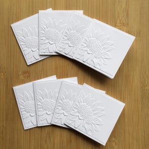May include: A set of ten blank white cards with an embossed sunflower design. The cards are arranged in two stacks of five.