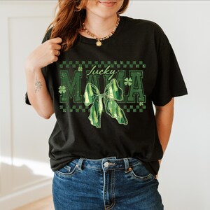 Peut inclure: T-shirt noir avec l'inscription "Lucky Mama" en vert, orn&eacute;e d'un n&oelig;ud vert et d'un motif de tr&egrave;fle. Le t-shirt a une coupe d&eacute;contract&eacute;e et est associ&eacute; &agrave; un jean bleu.