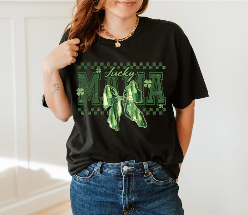 Peut inclure: T-shirt noir avec l'inscription "Lucky Mama" en vert, orn&eacute;e d'un n&oelig;ud vert et d'un motif de tr&egrave;fle. Le t-shirt a une coupe d&eacute;contract&eacute;e et est associ&eacute; &agrave; un jean bleu.