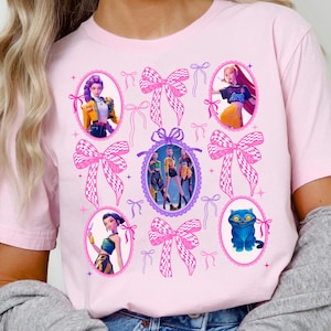 May include: A light pink t-shirt featuring a graphic print of multiple oval frames containing animated characters and a blue cat, interspersed with large pink and white chequered bows and smaller pink ribbon illustrations throughout the design.