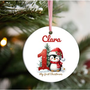 Puede incluir: Adorno navideño blanco con una cinta roja, con un pingüino con gorro y bufanda de Papá Noel. El adorno tiene el nombre "Clara", el número "1" y el texto "My first Christmas".