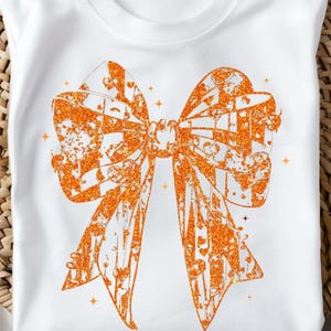 Pode incluir: Camiseta branca com um grande design de laço laranja brilhante. O laço tem um padrão texturizado com detalhes de corações e flores. A camiseta está dobrada sobre uma superfície tecida.