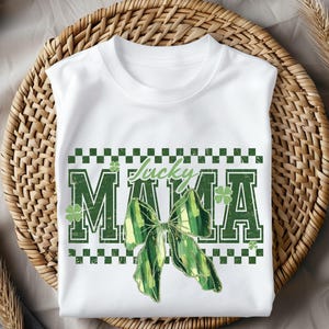 Peut inclure: T-shirt blanc avec un motif de n&oelig;ud vert et blanc et le texte "Lucky MAMA". Le design comprend des tr&egrave;fles verts et une bordure &agrave; carreaux, parfait pour la Saint-Patrick.