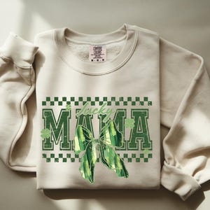 Peut inclure: Sweat-shirt cr&egrave;me avec l'inscription "Lucky MAMA" en vert, orn&eacute; d'un n&oelig;ud vert et de tr&egrave;fles. Le motif est encadr&eacute; par un motif &agrave; carreaux verts et blancs. Le sweat-shirt a des manches longues et un col rond.