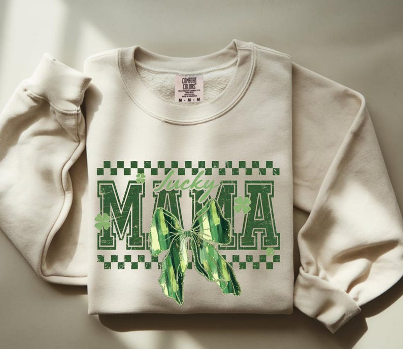 Peut inclure: Sweat-shirt cr&egrave;me avec l'inscription "Lucky MAMA" en vert, orn&eacute; d'un n&oelig;ud vert et de tr&egrave;fles. Le motif est encadr&eacute; par un motif &agrave; carreaux verts et blancs. Le sweat-shirt a des manches longues et un col rond.