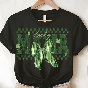 Peut inclure: T-shirt noir avec un motif de n&oelig;ud vert et blanc et les mots "Lucky Mama" en vert. Le motif comprend des tr&egrave;fles et un motif &agrave; carreaux, parfait pour la Saint-Patrick.