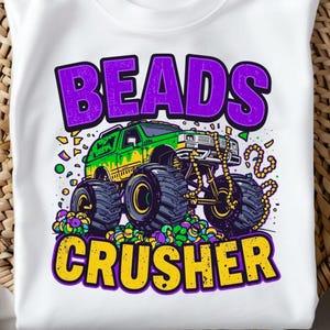 May include: White t-shirt featuring a colorful graphic of a monster truck with large tires, a green and yellow body, and the text "BEADS CRUSHER" in purple and yellow. The design includes Mardi Gras beads and confetti.
