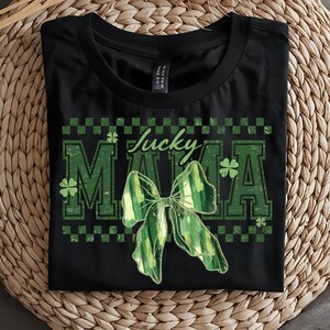 Peut inclure: T-shirt noir avec l'inscription "Lucky Mama" en vert, orn&eacute; d'un n&oelig;ud vert et de tr&egrave;fles. Le design a un aspect vieilli et vintage, parfait pour la Saint-Patrick.