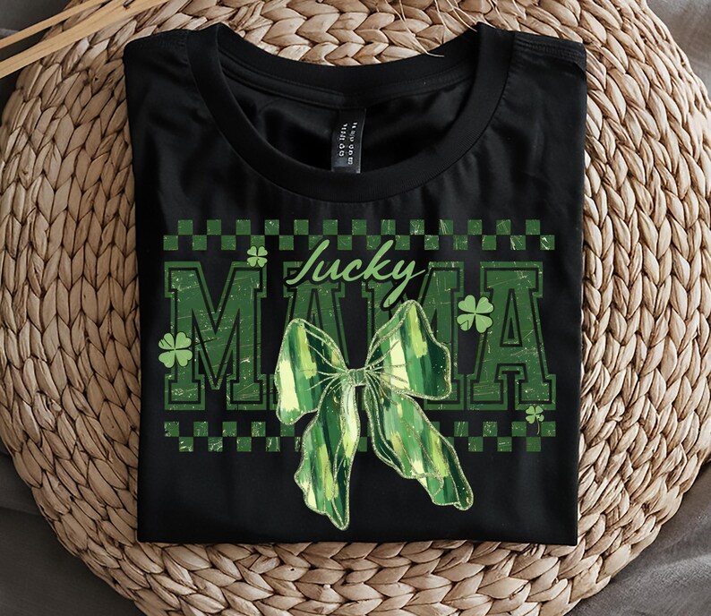 Peut inclure: T-shirt noir avec l'inscription "Lucky Mama" en vert, orn&eacute; d'un n&oelig;ud vert et de tr&egrave;fles. Le design a un aspect vieilli et vintage, parfait pour la Saint-Patrick.
