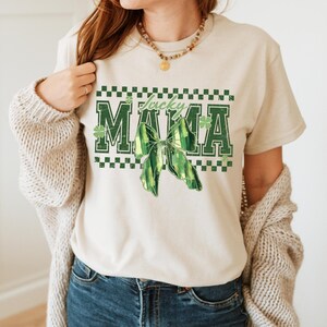 Peut inclure: Un t-shirt cr&egrave;me avec les mots "Lucky Mama" en lettres vertes, un n&oelig;ud vert et des motifs de tr&egrave;fles. Le t-shirt a un col rond et des manches courtes. Le motif est centr&eacute; sur le devant du t-shirt.