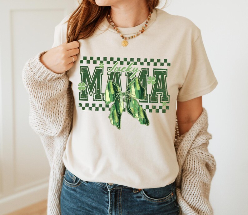 Peut inclure: Un t-shirt cr&egrave;me avec les mots "Lucky Mama" en lettres vertes, un n&oelig;ud vert et des motifs de tr&egrave;fles. Le t-shirt a un col rond et des manches courtes. Le motif est centr&eacute; sur le devant du t-shirt.