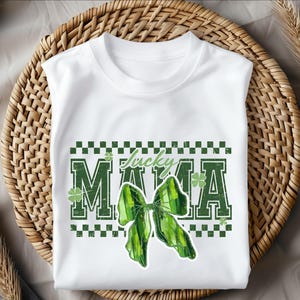 Peut inclure: T-shirt blanc avec un motif vert et blanc. Le t-shirt pr&eacute;sente les mots "Lucky Mama" dans une police verte vieillie, un n&oelig;ud vert et des motifs de tr&egrave;fles. Le motif est plac&eacute; sur une bordure &agrave; carreaux.