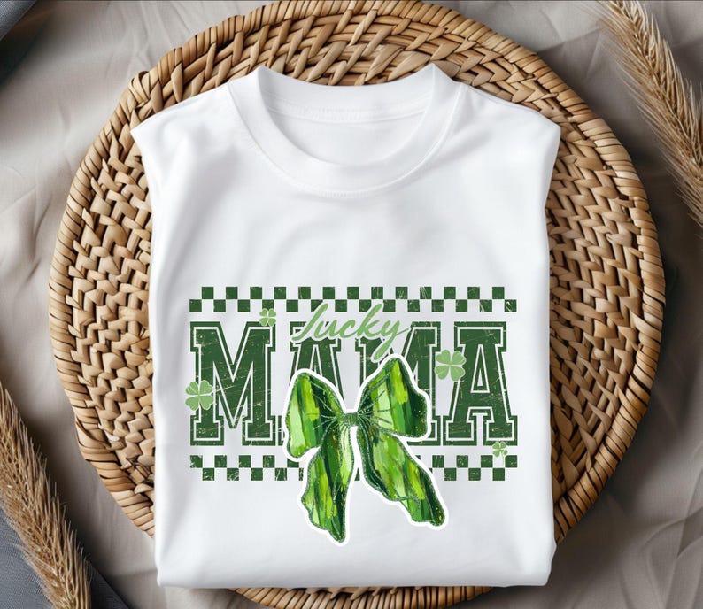 Peut inclure: T-shirt blanc avec un motif vert et blanc. Le t-shirt pr&eacute;sente les mots "Lucky Mama" dans une police verte vieillie, un n&oelig;ud vert et des motifs de tr&egrave;fles. Le motif est plac&eacute; sur une bordure &agrave; carreaux.