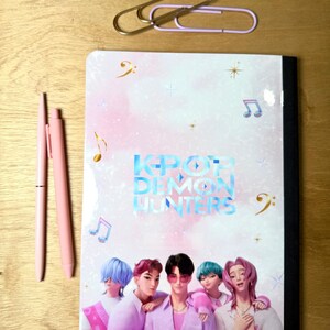 May include: A notebook with the text "KPOP DEMON HUNTERS" and an illustration of five people in pink and purple clothing. Two pink pens and two paper clips are also on the wooden surface.