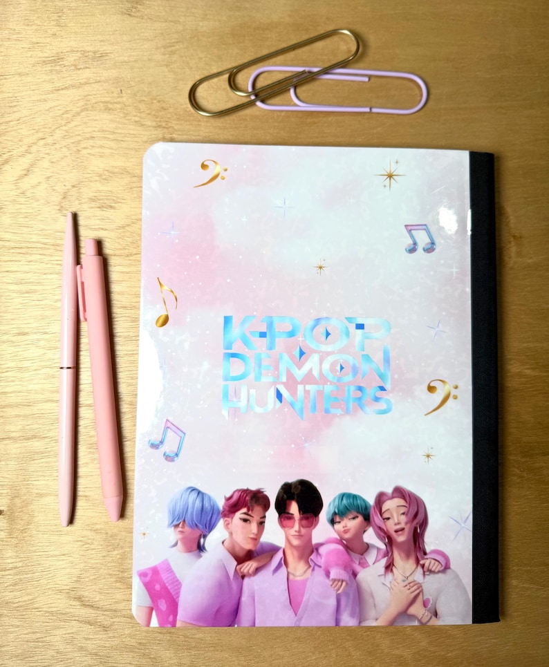 May include: A notebook with the text "KPOP DEMON HUNTERS" and an illustration of five people in pink and purple clothing. Two pink pens and two paper clips are also on the wooden surface.