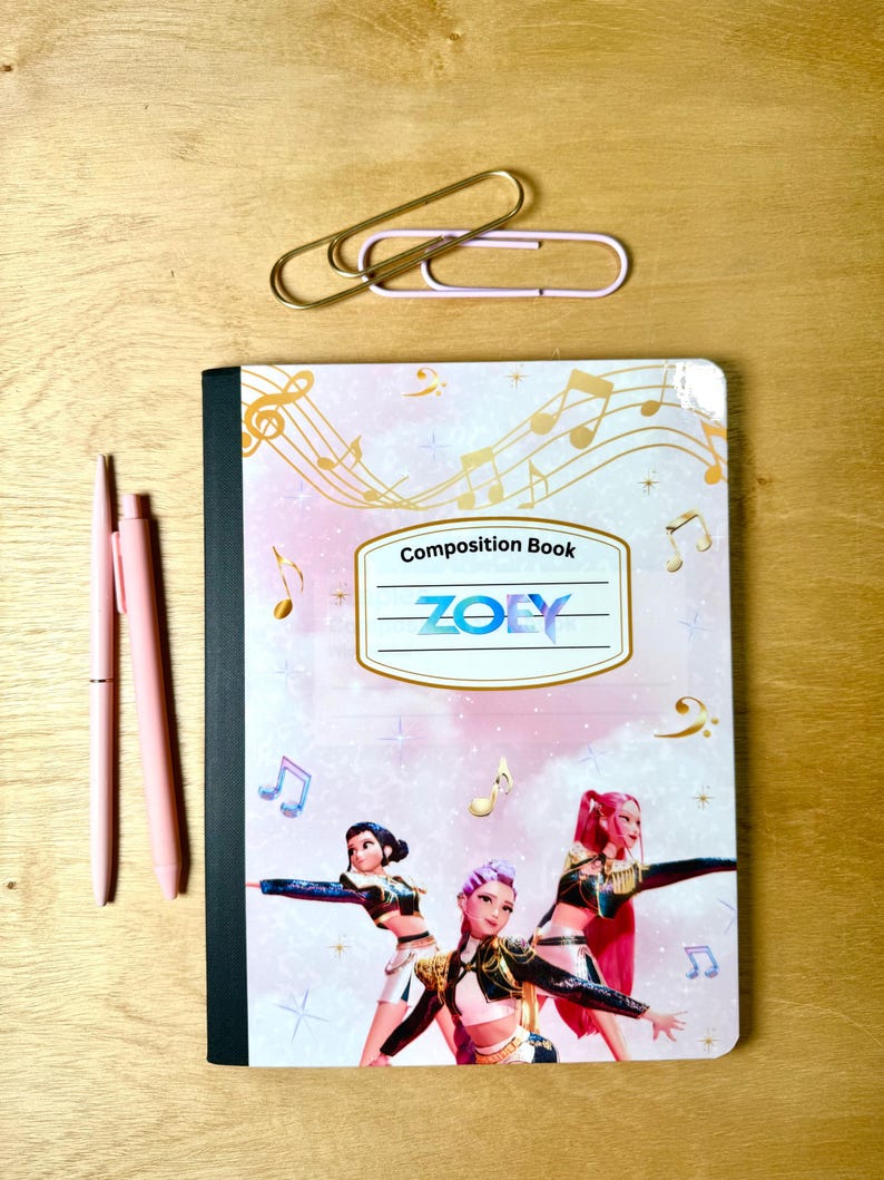 May include: A composition book with a musical theme, featuring a design with musical notes and the text "ZOEY". Two pink pens and two paper clips are next to the notebook on a wooden surface.