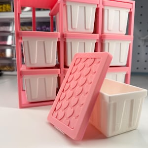 May include: A pink storage organizer with nine white and pink bins. A pink lid with heart-shaped cutouts rests beside an open bin. The organizer is designed to hold small items, perfect for craft supplies or small tools.